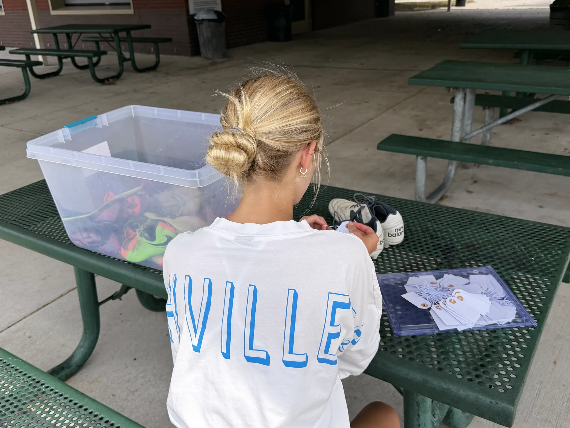 A volunteer at a picnic table beside a donation bin, sorting cleats into pairs and writing size tags